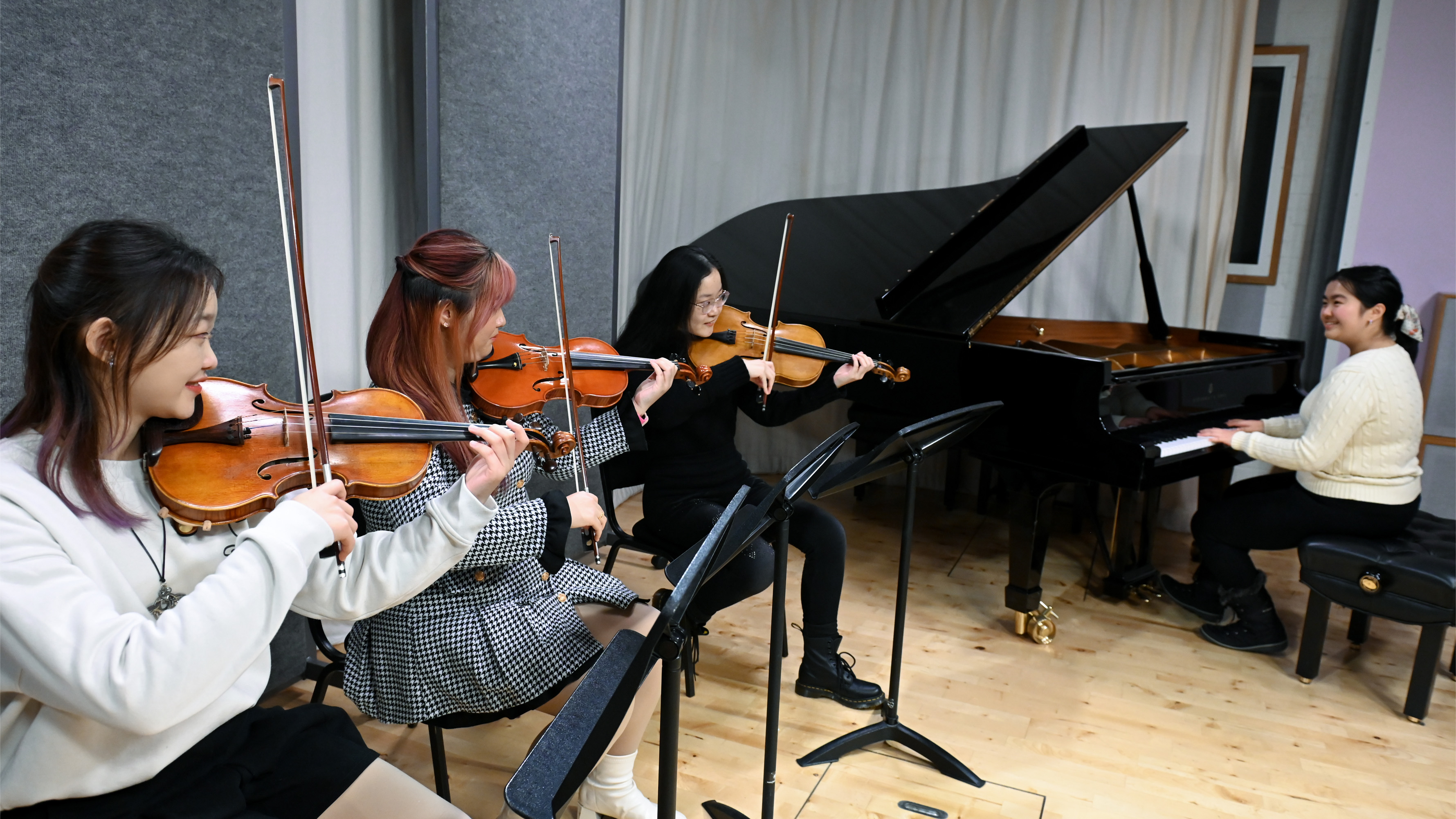 Three female students performing on violins, with another female student accompanying on piano, smiling at each other.